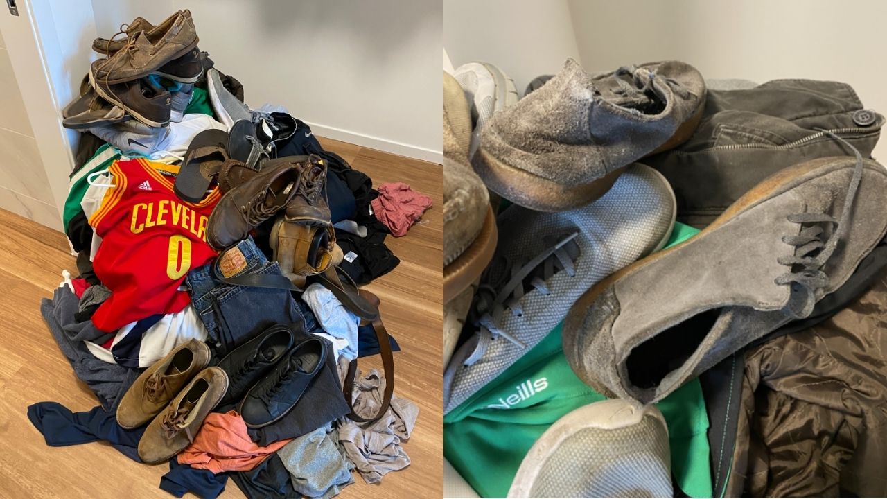 A pile of assorted clothing and shoes, including sneakers, dress shoes, and a red Cleveland basketball jersey, scattered on a wooden floor.